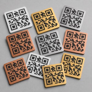 Op de afbeelding: Een verzameling vierkante metalen onderzetters met QR-code ontwerpen. De onderzetters zijn er in verschillende metallic afwerkingen, waaronder zilver, goud en koper. De QR-codes zijn zwart.