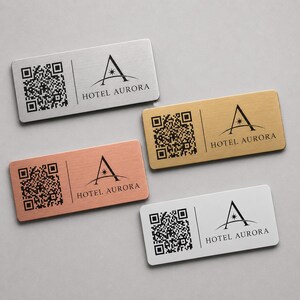 May include: Four rectangular metal name tags in silver, gold, copper, and white. Each tag features a QR code on the left and the "HOTEL AURORA" logo on the right. The logo is a stylized "A" with a star.