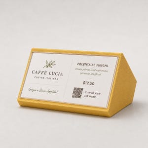 Puede incluir: Un expositor de mesa amarillo con el texto "CAFFÈ LUCIA CUCINA ITALIANA" y "POLENTA AL FUNGHI" con una descripción del plato. Incluye un código QR y la frase "SCAN TO VIEW OUR MENU".