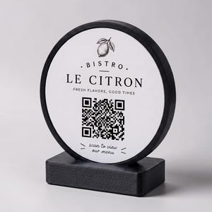 Op de afbeelding: Een zwart-wit bistro-bord met de tekst ". BISTRO. LE CITRON FRESH FLAVORS, GOOD TIMES" en een QR-code. Het bord is rond en gemonteerd op een rechthoekige basis.