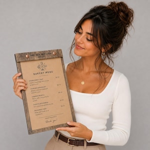 Op de afbeelding: Een houten bakkerijmenu met de tekst "THE OLD RED LION EST. 1577" bovenaan. Het menu bevat bakkerijartikelen zoals zuurdesembrood, amandelcroissant, kaneelbroodje, chocolade barka en seizoensgebonden taart. Het menu wordt door een persoon vastgehouden.