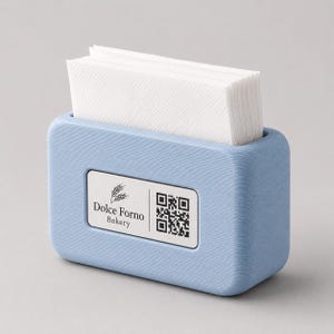 Op de afbeelding: Een lichtblauwe servethouder met een stapel witte servetten. De houder heeft een textuuroppervlak en een rechthoekig label met de tekst "Dolce Forno Bakery" en een QR-code.
