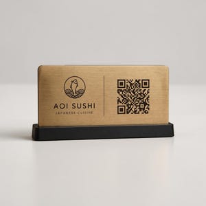 Op de afbeelding: Een rechthoekig bord in goudkleur met de tekst "AOI SUSHI JAPANESE CUISINE" en een QR-code. Het bord is gemonteerd op een zwarte voet. Het logo toont een vis boven golven.