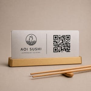 Könnte beinhalten: Ein rechteckiges silbernes Schild mit dem Text "AOI SUSHI JAPANESE CUISINE" und einem QR-Code. Das Schild steht auf einem goldfarbenen Sockel, mit einem Paar Essstäbchen davor.