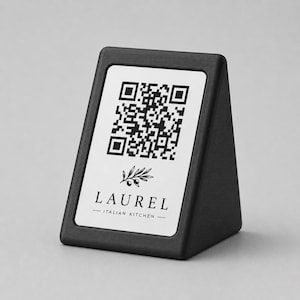 Könnte beinhalten: Ein schwarzer, dreieckiger Tischaufsteller mit einem weißen Einsatz, der einen schwarzen QR-Code zeigt. Unter dem Code befindet sich eine kleine Illustration eines Olivenzweigs sowie der Text LAUREL ITALIAN KITCHEN in schwarzer Serifenschrift.