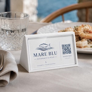 Puede incluir: Un expositor de mesa blanco con el texto "MARE BLU - RISTORANTE DI PESCE - AUTHENTIC ITALIAN SEAFOOD" y un logotipo de pez. Un código QR está a la derecha con el texto "Scan for menu". El expositor está sobre una mesa con un vaso de agua y un plato de comida al fondo.