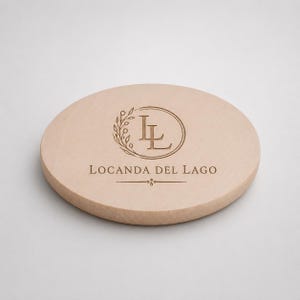 Pode incluir: Um porta-copos redondo de madeira clara com um logótipo gravado em castanho escuro. O logótipo consiste nas letras LL dentro de uma moldura circular com um ramo de folhas no lado esquerdo, seguido do texto LOCANDA DEL LAGO e uma linha.