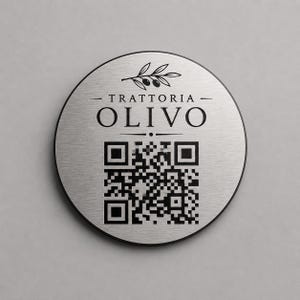 Op de afbeelding: Een ronde, geborstelde metalen plaat met een zwarte rand. De plaat toont de tekst "- TRATTORIA - OLIVO" in een vetgedrukte letter, een QR-code en een olijftakillustratie. De achtergrond is lichtgrijs.