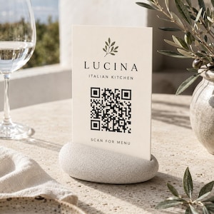 Pode incluir: Um cartão de menu retangular na cor creme com um ícone de folha preta, o texto LUCINA ITALIAN KITCHEN, um código QR e o texto SCAN FOR MENU. O cartão é mantido na vertical por uma base de pedra oval, cinza-clara e com superfície texturizada.
