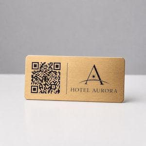 May include: A rectangular, brushed gold name tag with rounded corners. The tag features a QR code on the left and the words "HOTEL AURORA" below a stylized "A" logo on the right. The logo includes a star.