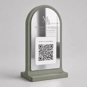 Pode incluir: Um sinal de mesa arqueado com uma moldura verde sálvia e uma superfície espelhada. O espelho apresenta o texto MAISON DORÉE CUISINE & COCKTAILS e uma inserção quadrada branca exibindo um código QR preto com o texto SCAN TO VIEW MENU.