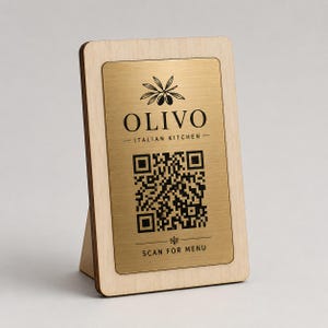 Puede incluir: Un soporte rectangular de madera con una placa de metal dorado cepillado con texto negro y un código QR. La placa muestra un gráfico de rama de olivo, el texto OLIVO ITALIAN KITCHEN y la instrucción SCAN FOR MENU en la parte inferior.