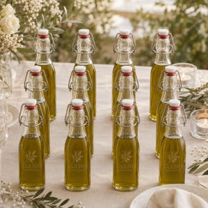 Può includere: Una collezione di dodici bottiglie in vetro trasparente riempite di olio d'oliva dorato, ciascuna con un tappo meccanico con chiusura bianca e guarnizione rossa. Ogni bottiglia è incisa con un ramo d'ulivo e il testo L'ULIVO RISTORANTE.