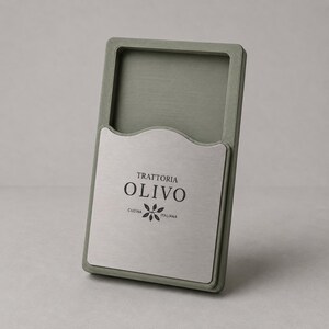 Op de afbeelding: Een rechthoekige, saliegroene menuhouder voor restaurants met een geborstelde metalen inzet. De inzet toont de tekst "TRATTORIA OLIVO" in een schreefletter, met daaronder "CUCINA ITALIANA". Een gestileerde olijftakafbeelding is ook aanwezig.