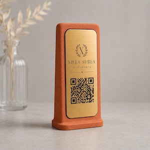 Può includere: Un cartello del ristorante color terracotta con una placca dorata. La placca presenta il testo "VILLA AUREA RISTORANTE" e un codice QR. Il cartello è rettangolare con una base larga.