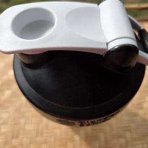 K&ouml;nnte beinhalten: Eine schwarze Wasserflasche mit wei&szlig;em Deckel und Griff. Der Deckel hat eine kreisf&ouml;rmige &Ouml;ffnung und einen Klappverschluss. Die Flasche scheint aus Kunststoff zu sein und ist f&uuml;r Sport oder Outdoor-Aktivit&auml;ten konzipiert.