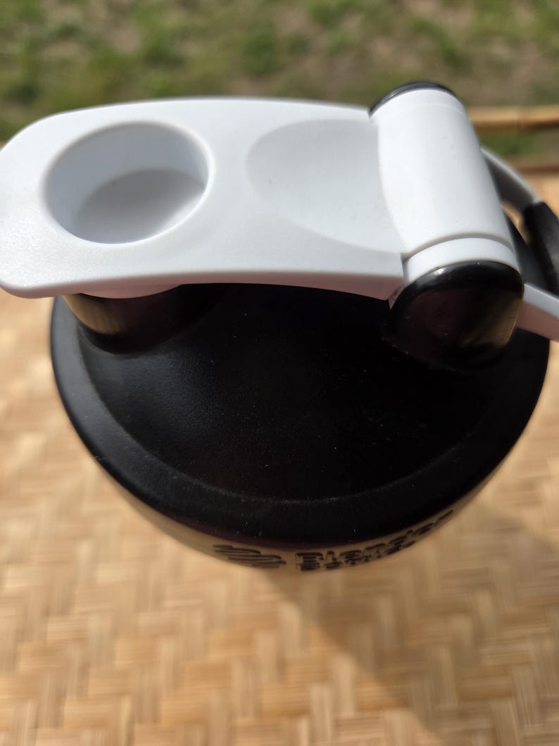 K&ouml;nnte beinhalten: Eine schwarze Wasserflasche mit wei&szlig;em Deckel und Griff. Der Deckel hat eine kreisf&ouml;rmige &Ouml;ffnung und einen Klappverschluss. Die Flasche scheint aus Kunststoff zu sein und ist f&uuml;r Sport oder Outdoor-Aktivit&auml;ten konzipiert.