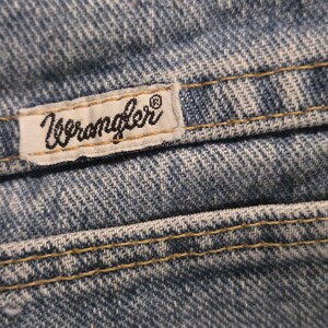Może przedstawiać: Zbliżenie niebieskich dżins&oacute;w z widocznymi szwami i białą metką. Etykieta zawiera czarne logo "Wrangler". Dżinsy mają klasyczny, zużyty wygląd, ze szczeg&oacute;łowymi szwami i teksturowaną tkaniną.