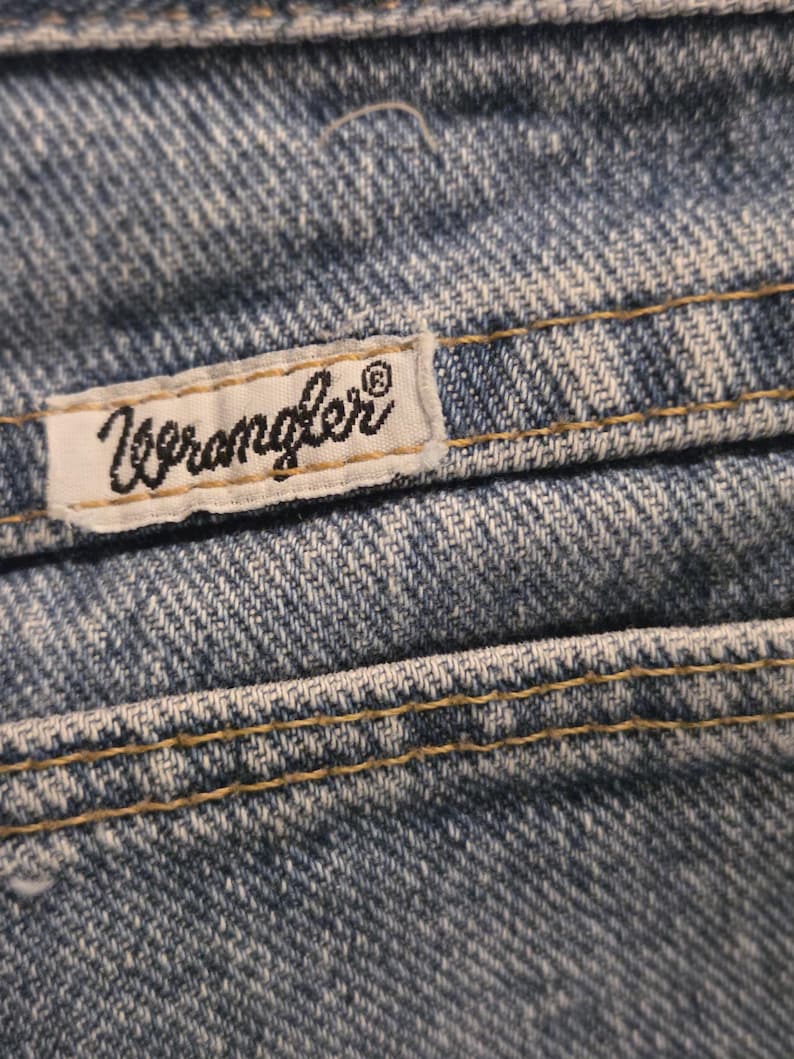 Może przedstawiać: Zbliżenie niebieskich dżins&oacute;w z widocznymi szwami i białą metką. Etykieta zawiera czarne logo "Wrangler". Dżinsy mają klasyczny, zużyty wygląd, ze szczeg&oacute;łowymi szwami i teksturowaną tkaniną.