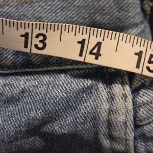 Może przedstawiać: Zbliżenie jasnoniebieskich dżins&oacute;w z miarką. Taśma pokazuje pomiary od 33 do 38 cm. Dżinsy mają widoczne szwy i klasyczną fakturę dżinsu.