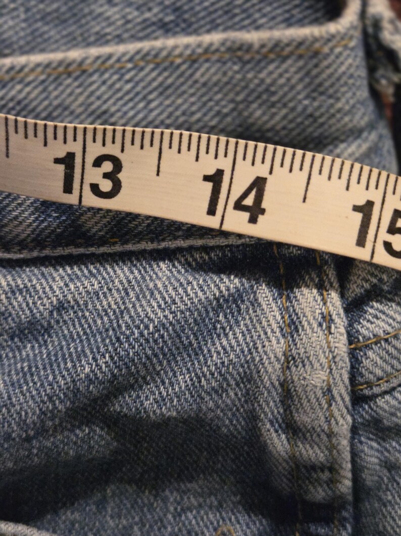 Może przedstawiać: Zbliżenie jasnoniebieskich dżins&oacute;w z miarką. Taśma pokazuje pomiary od 33 do 38 cm. Dżinsy mają widoczne szwy i klasyczną fakturę dżinsu.