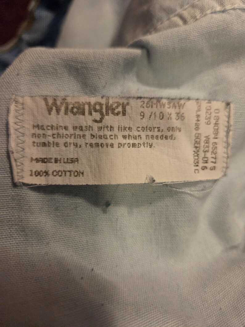 Może przedstawiać: Zbliżenie na jasnoniebieską tkaninę z białą etykietą. Etykieta głosi "Wrangler" i zawiera instrukcje prania oraz szczeg&oacute;ły dotyczące rozmiaru: 26HWSAW 9/10 x 36. Tkanina jest w 100% bawełniana i wyprodukowana w USA.