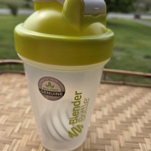 May include: A clear Blender Bottle with a lime green lid and accents. The bottle has a white flip-top closure and a stainless steel mixing ball inside. The bottle is BPA-free and has the Blender Bottle logo printed on the side.