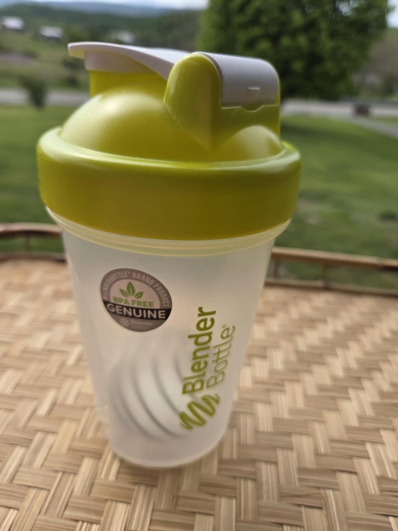 May include: A clear Blender Bottle with a lime green lid and accents. The bottle has a white flip-top closure and a stainless steel mixing ball inside. The bottle is BPA-free and has the Blender Bottle logo printed on the side.