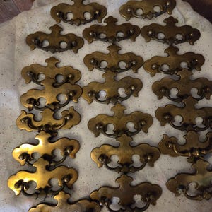 May include: A collection of antique brass drawer pulls. Each pull features a decorative design with a curved handle and ornate detailing. The pulls are arranged on a white surface, showcasing their aged, golden color. Ideal for vintage furniture restoration.