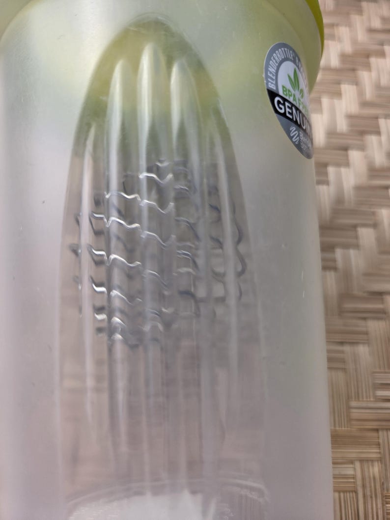 May include: A clear plastic blender bottle with a green lid and a stainless steel whisk. The bottle has a textured design and a BPA-free label. The bottle is designed for mixing protein shakes and other beverages.