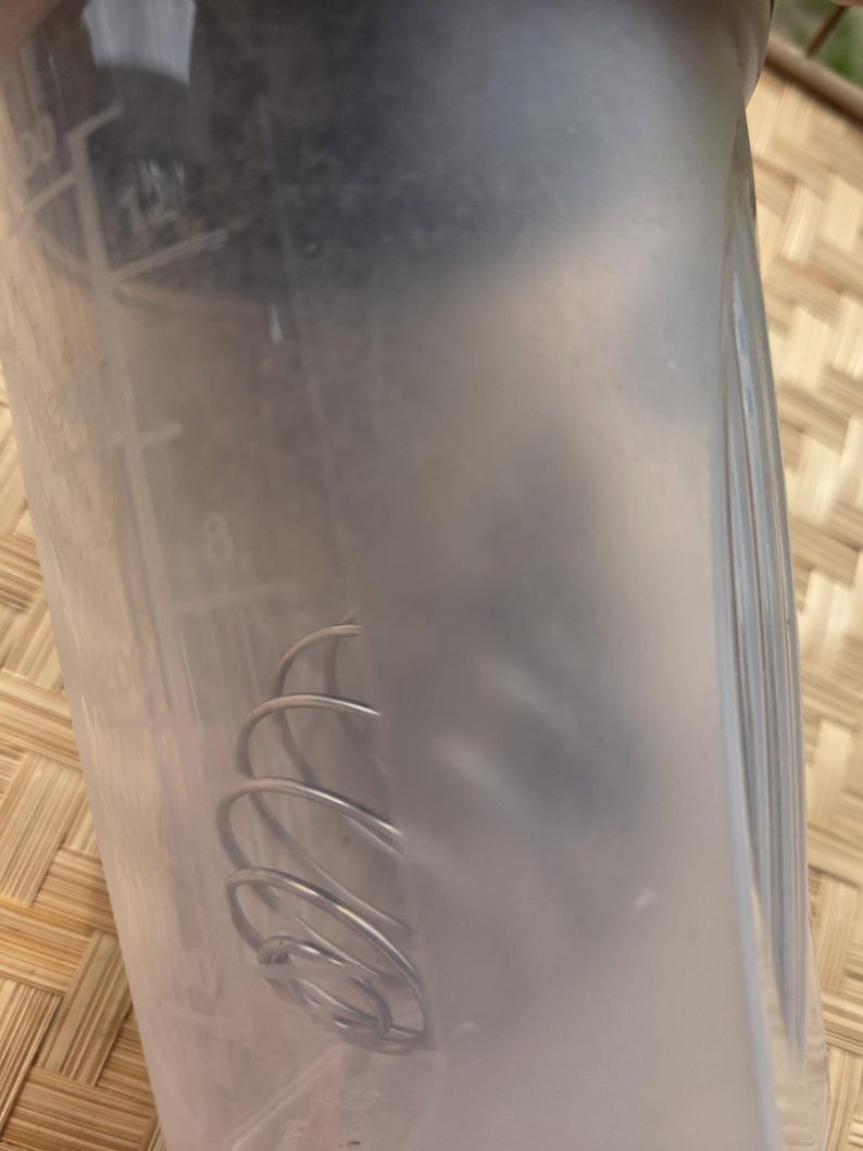 K&ouml;nnte beinhalten: Eine transparente Kunststoff-Shakerflasche mit Messmarkierungen. Im Inneren der Flasche befindet sich ein silberner Drahtquirl. Die Flasche ist teilweise mit einer tr&uuml;ben Fl&uuml;ssigkeit gef&uuml;llt. Der Hintergrund ist eine Oberfl&auml;che aus geflochtenem Bambus.