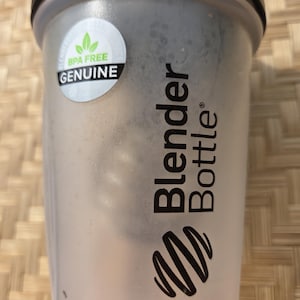 K&ouml;nnte beinhalten: Eine transparente Blender Bottle mit schwarzem Deckel und einem wei&szlig;en Aufkleber mit der Aufschrift "BPA FREE GENUINE". Die Flasche hat das Blender Bottle Logo und Text in Schwarz.