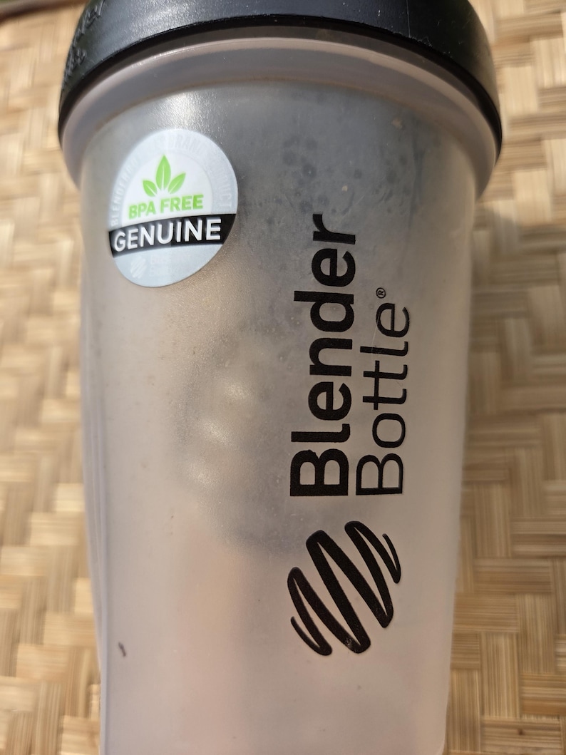 K&ouml;nnte beinhalten: Eine transparente Blender Bottle mit schwarzem Deckel und einem wei&szlig;en Aufkleber mit der Aufschrift "BPA FREE GENUINE". Die Flasche hat das Blender Bottle Logo und Text in Schwarz.