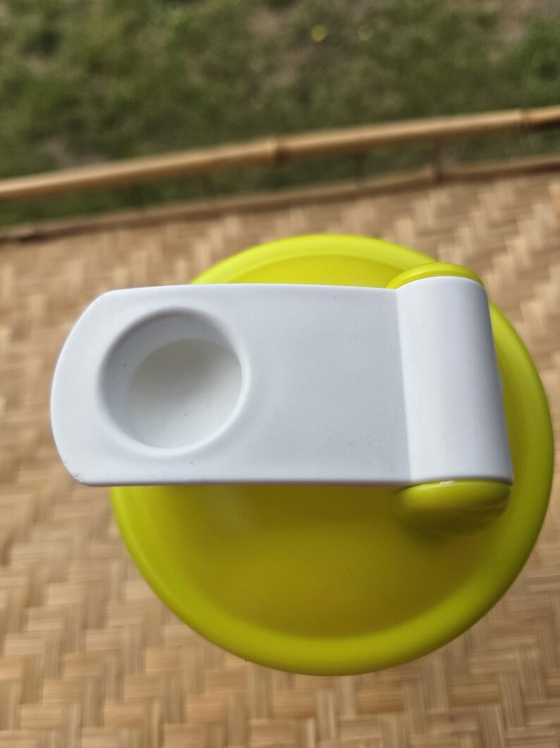 May include: A close-up of a yellow and white water bottle lid. The lid is bright yellow with a white flip-top closure. The closure has a circular opening. The background is a woven bamboo mat.
