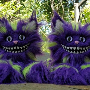 May include: Two plush Cheshire Cat slippers. The slippers are purple with green and white accents. They have large, smiling mouths with white teeth, pink noses, and large, round eyes. The slippers are designed to look like the Cheshire Cat from Alice in Wonderland.
