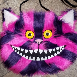Op de afbeelding: Een roze en paarse harige Cheshire Cat tas met een brede grijns en gele ogen.