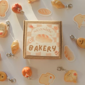 Pode incluir: Uma coleção de chaveiros e adesivos em miniatura com tema de comida. Os chaveiros incluem pão, peixe e outros produtos de panificação. Uma pequena caixa com as palavras "Seaside Bakery" está no centro, com um fundo quadriculado.