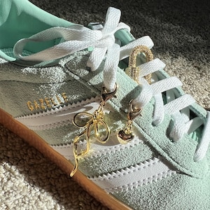 May include: A pair of light green suede sneakers with white laces and white stripes. The laces are adorned with gold charms, including a heart, a bow, and a padlock.