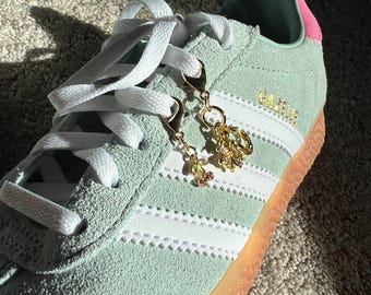 Może przedstawiać: Para jasnozielonych skórzanych butów Adidas Gazelle z białymi paskami i gumową podeszwą. Sznurowadła są zawiązane, a do nich przyczepione są złote breloki.