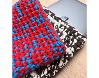 May include: Two crocheted laptop sleeves, one in red and blue stripes and the other in black and white stripes, are stacked on top of each other on a wooden surface.