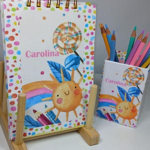 Pode incluir: Um conjunto colorido de bloco de notas e porta-lápis com um design de arco-íris, sol e pirulito. O bloco de notas tem uma encadernação em espiral e o porta-lápis tem uma ranura para lápis. Ambos os itens apresentam o nome "Carolina" em letras cor-de-rosa.