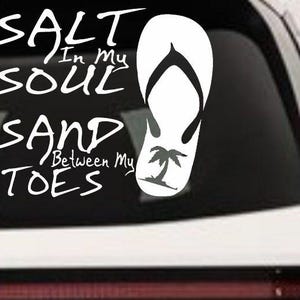 May include: White decal with the text "SALT IN MY SOUL SAND BETWEEN MY TOES" and two white flip-flops with palm tree graphics. The decal is applied to a car window.