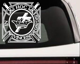 May include: A white vinyl decal featuring a cross and crown emblem centered within a cross-shaped frame. The text IN HOC SIGNO VINCES is arranged around the central circle, with two crossed swords positioned behind the upper arms of the cross.