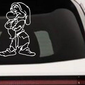 May include: A white vinyl decal featuring a line art illustration of a character with a large nose, a pointed hat, and crossed arms, standing in a pose with a grumpy expression. The design is applied to the rear window of a white vehicle.