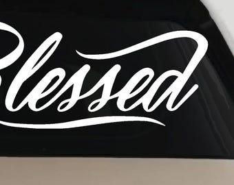 May include: A white vinyl decal featuring the word Blessed in a cursive script font. The lettering has a flowing, calligraphic style with an elongated tail on the letter d that underlines the word. The decal is applied to a dark surface.
