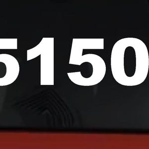 May include: A white vinyl decal featuring the text #5150 in a bold, sans-serif font. The decal is applied to the dark tinted glass of a vehicle window, with a portion of the red vehicle body visible at the bottom of the frame.