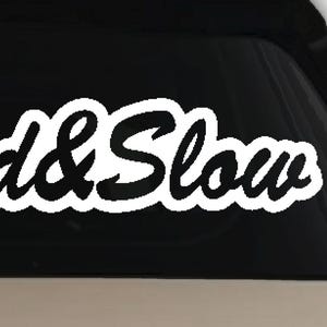 May include: A white vinyl decal sticker applied to a dark glass surface. The design features a stylized crown icon positioned above the text "Loud&Slow" written in a bold, cursive, script-style font with a thick white outline and black lettering.
