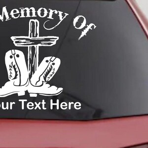 May include: A white vinyl decal for a vehicle window featuring the text In Memory Of arched over a wooden cross and a pair of cowboy boots, with the placeholder text Your Text Here positioned at the bottom of the graphic design.