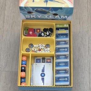 Puede incluir: La caja del juego de mesa Sky Team está abierta, revelando un inserto de plástico amarillo con compartimentos organizados. Contiene dados naranjas y azules, pequeñas fichas de avión blancas, marcadores rojos y negros, y cartas azules numeradas.