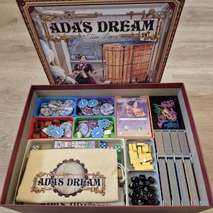 May include: The board game "Ada's Dream" is displayed open, revealing game pieces such as cards, tokens, and dice, neatly arranged in trays. The box lid features an illustration of a woman and a complex machine. The game's components are in a red box.